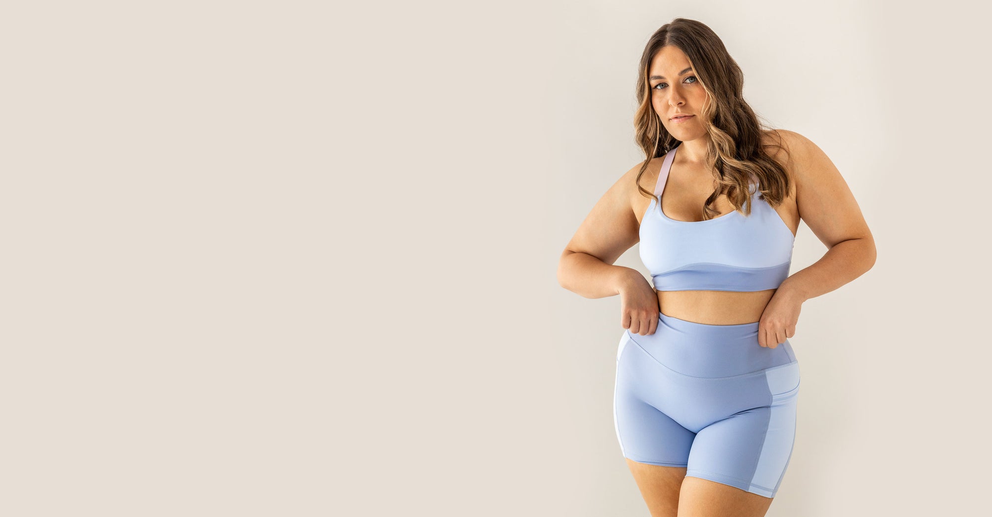 A curvy Earthletica Model wearing the Cascade Crop & Crystal Clear Pocket Set. She is looking directly at the camera, standing with her legs slightly crossed and her hands on the waistband of the shorts.