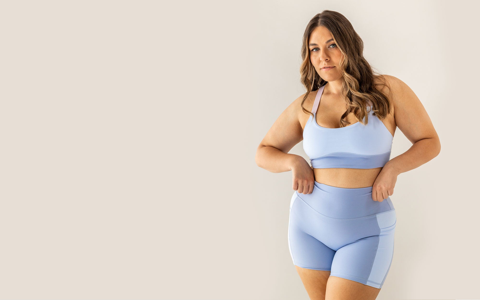 A curvy Earthletica Model wearing the Cascade Crop & Crystal Clear Pocket Set. She is looking directly at the camera, standing with her legs slightly crossed and her hands on the waistband of the shorts.