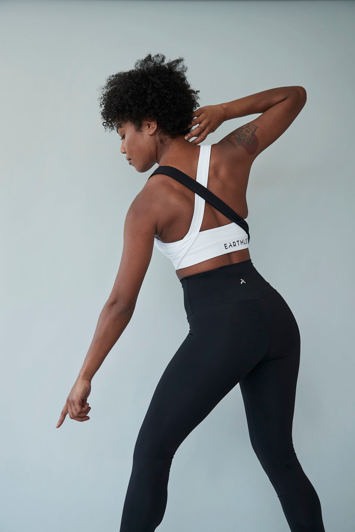 An Earthletica model photographed from behind wearing the Starry Night Crop + Midnight Leggings. She is in an active body pose with one arm raised and another pointing towards the floor.