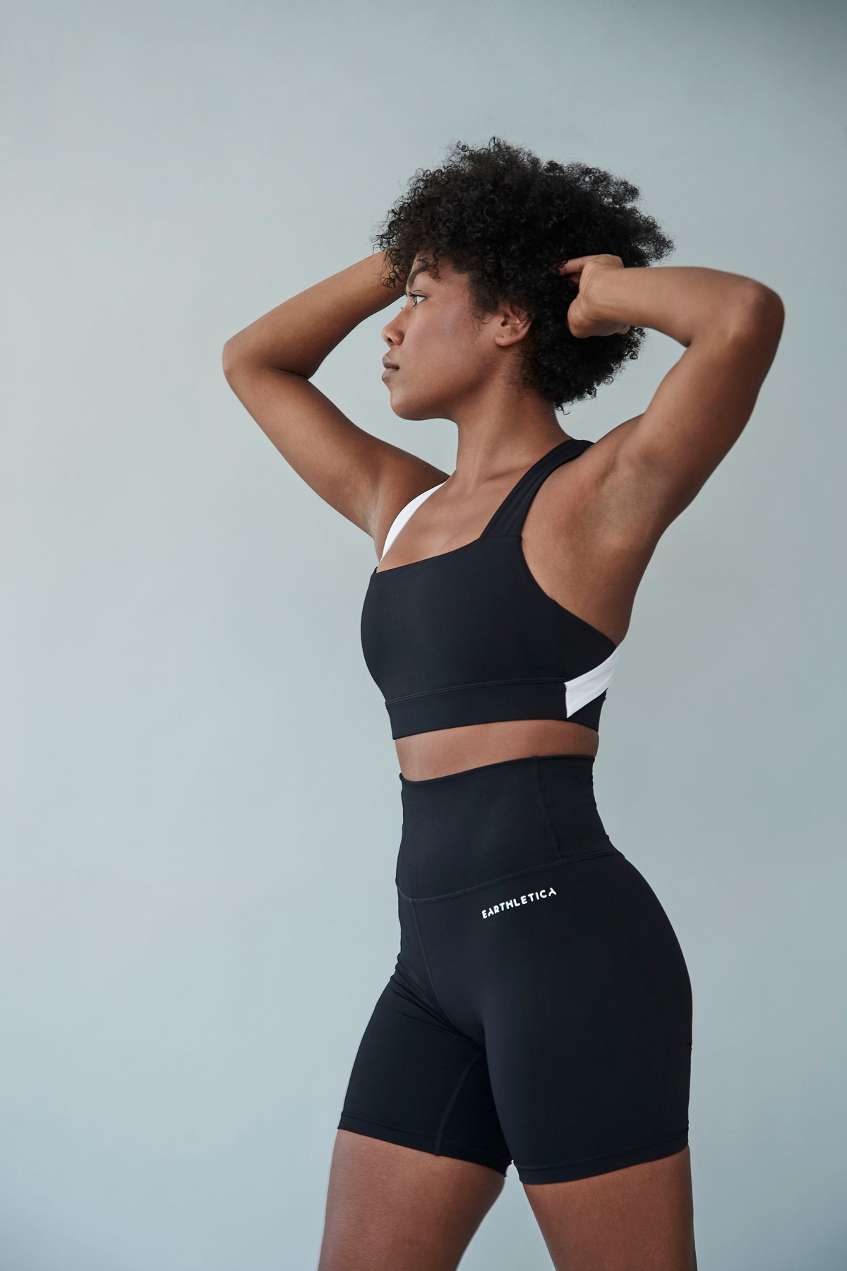 An Earthletica model wearing the Star Drop Crop + Midnight Bike Shorts activewear set. She is photographed from the side, with her arms raised above her head.