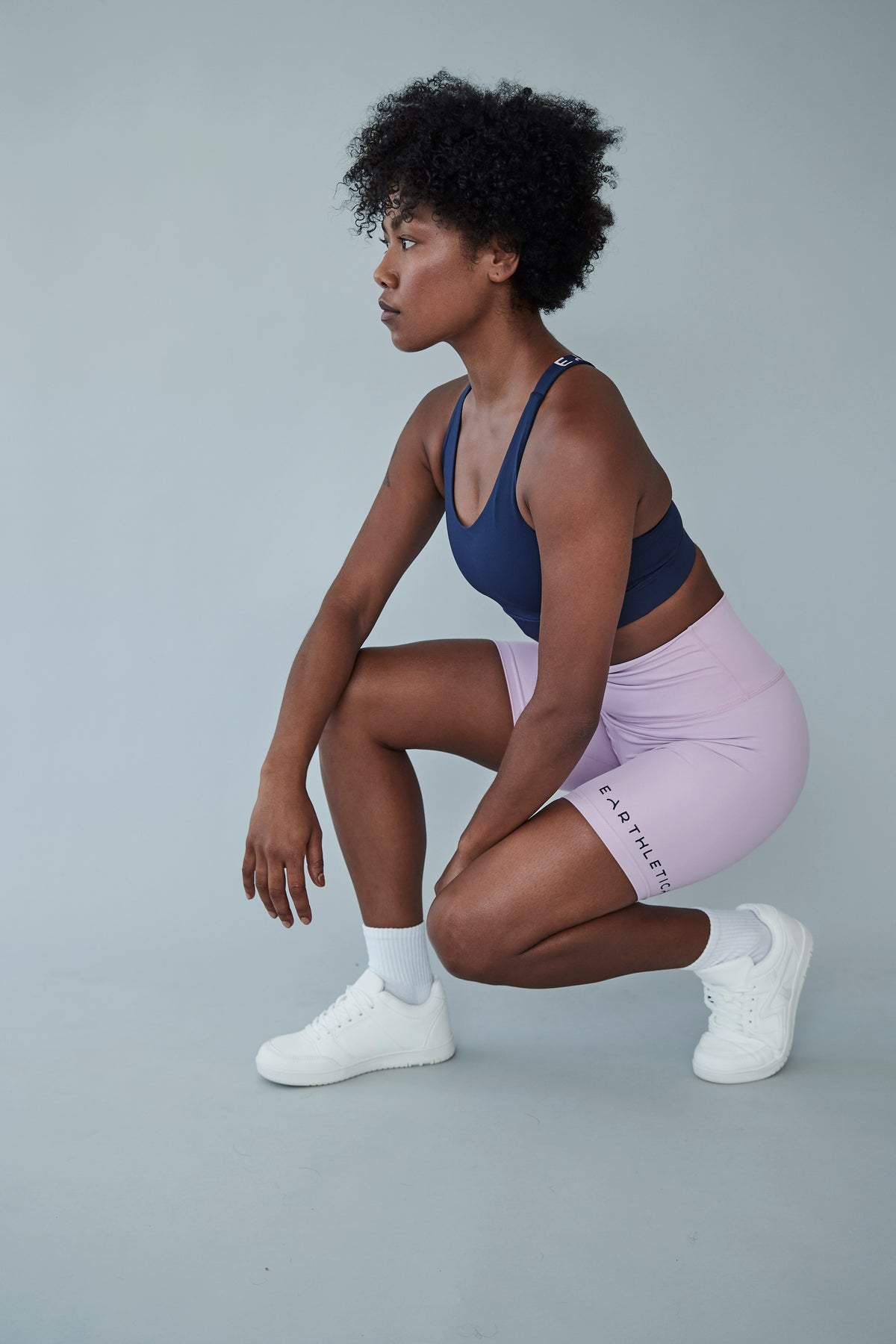An Earthletica model wearing the Into The Blue crop and Lilac Dreaming bike shorts. She is squatting down on one knee and looking off into the distance.
